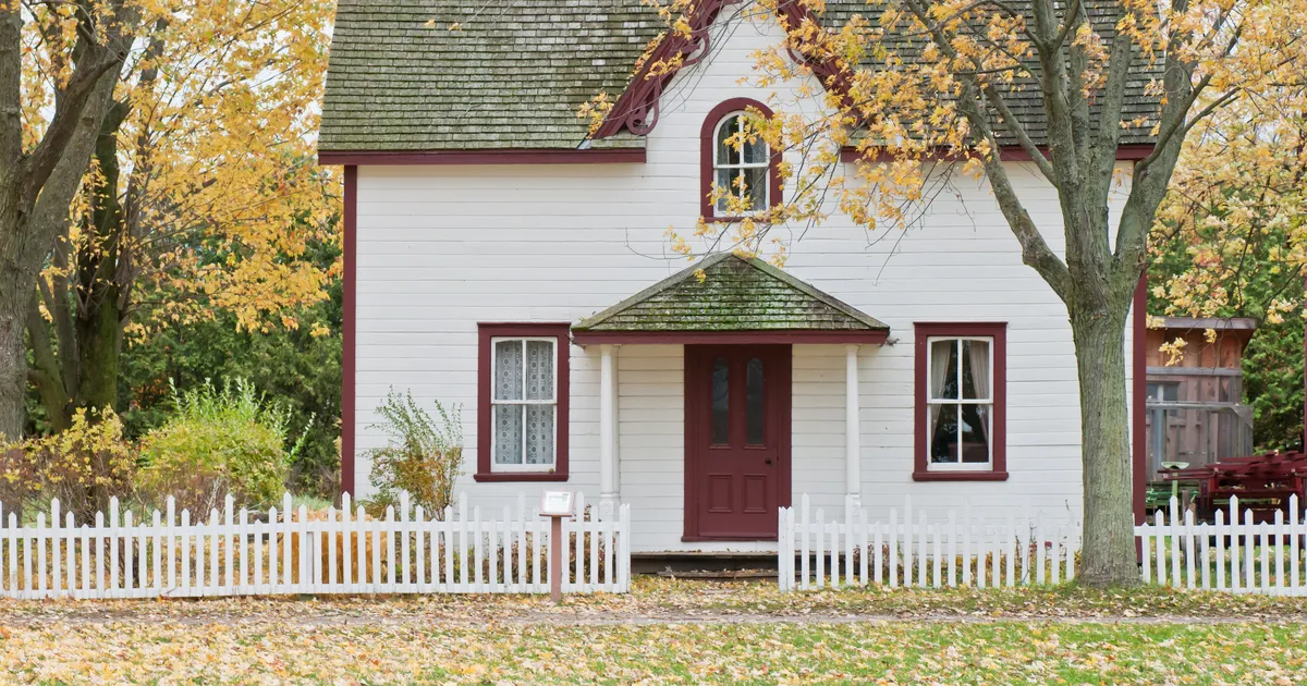 A white house with red trim behind a picket fence, with yellow fall trees overhead and leaves covering the front lawn